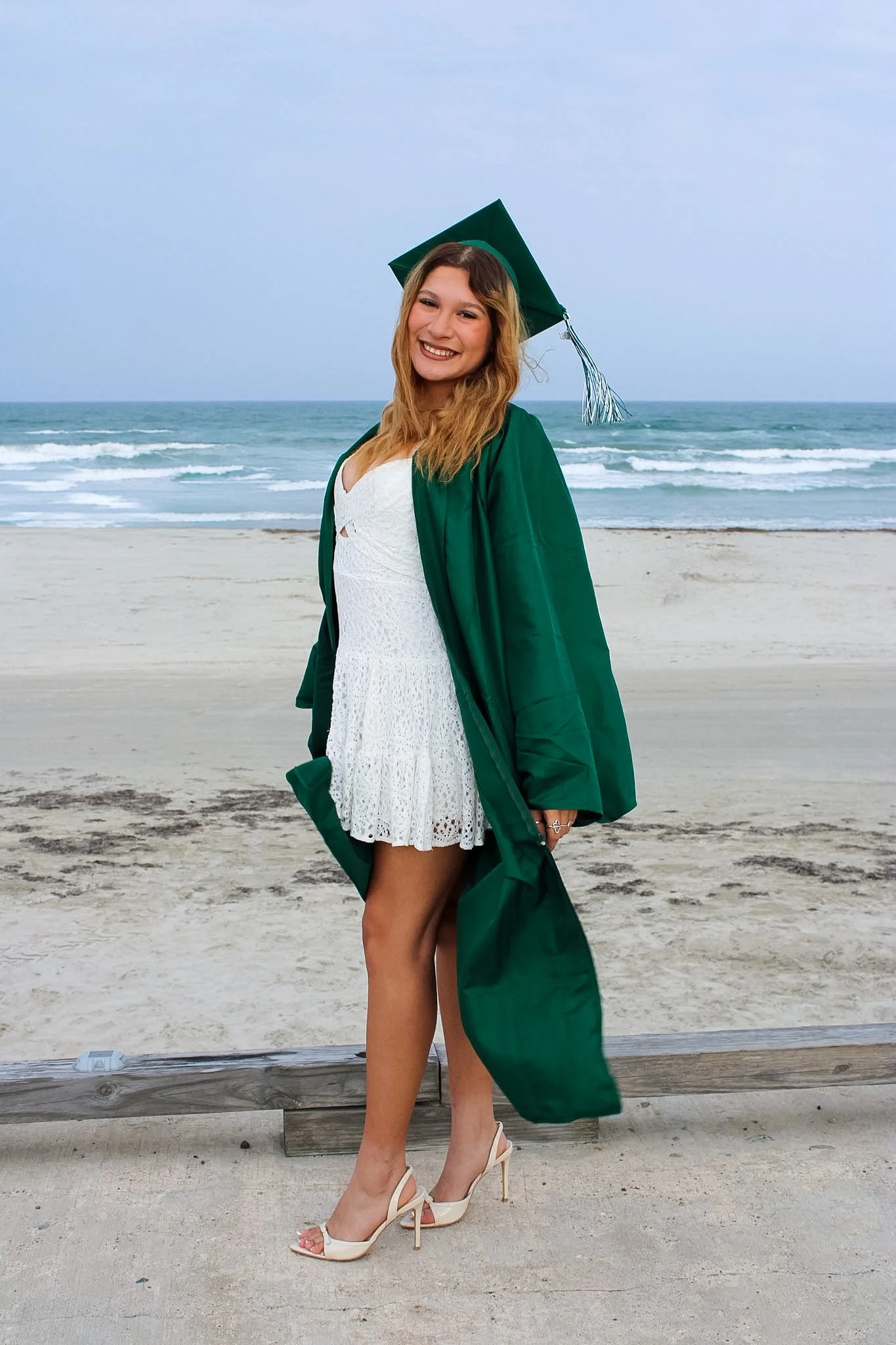 A young woman in a white dress, beige high heels, and a green graduation gown and cap standing on a beach with ocean waves in the background, smiling at the camera.