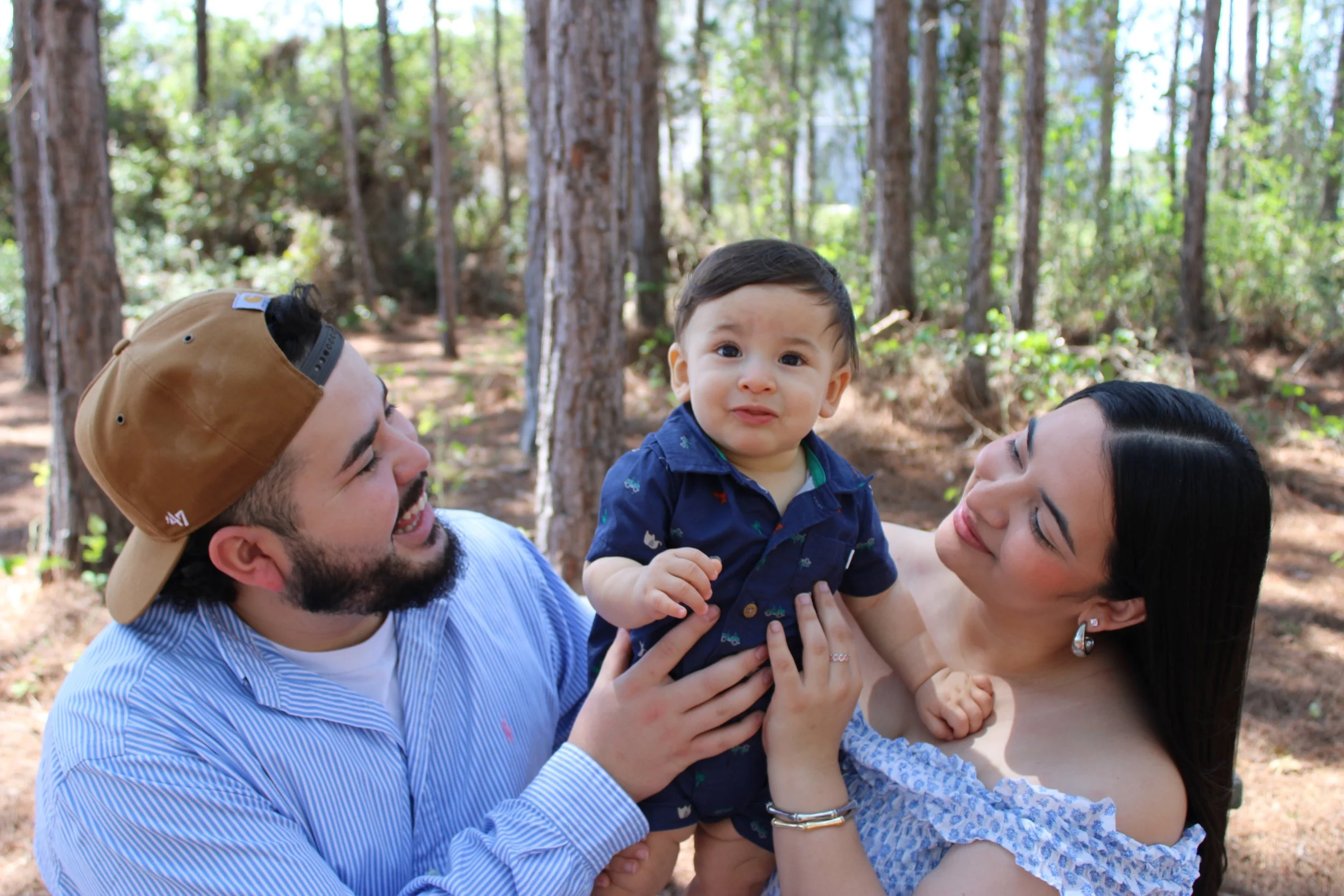 A family of three enjoying time together outdoors in a forest, with the father and mother holding their young son, all smiling and happy.