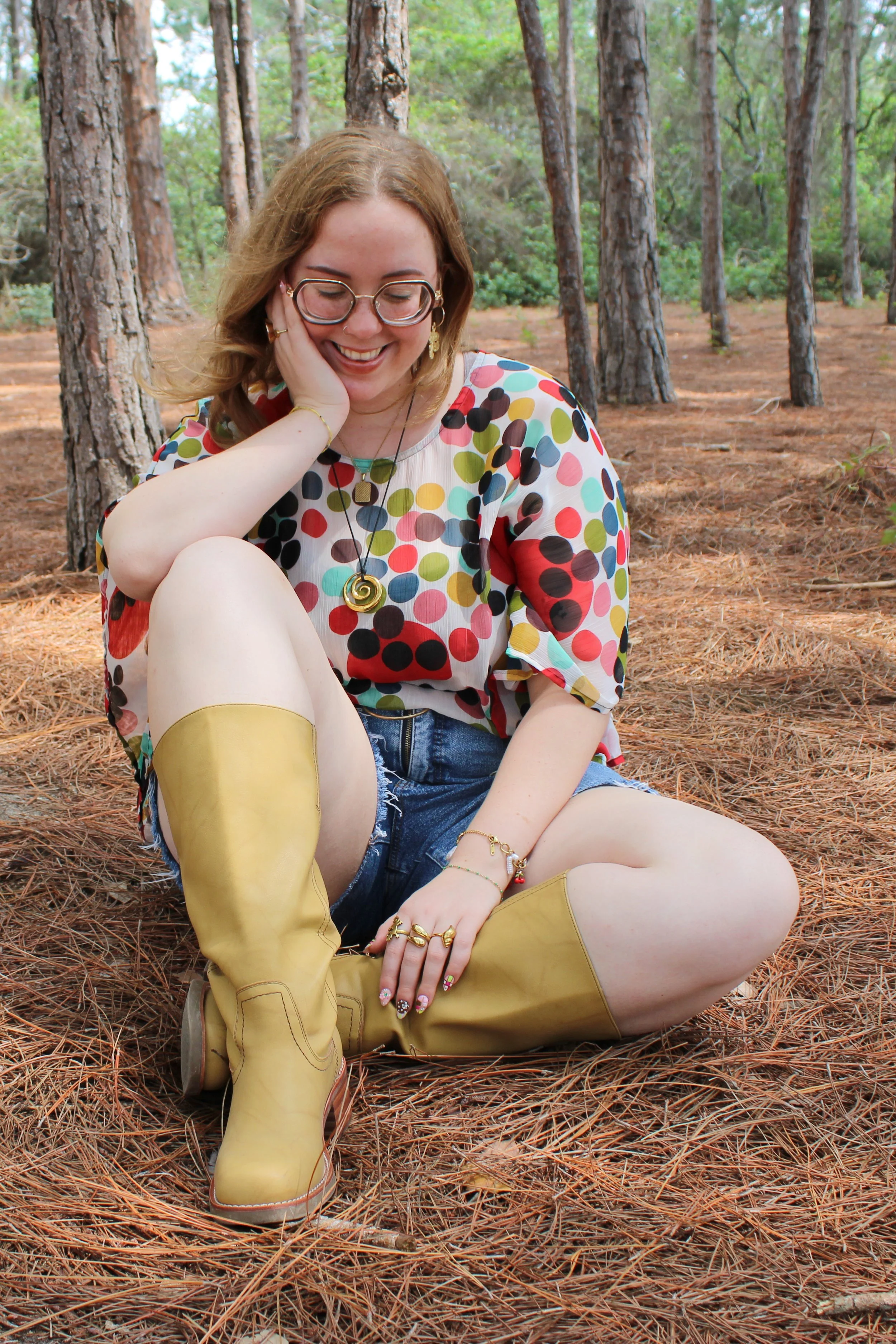 A young woman with light skin and glasses sitting on a forest ground covered with pine needles. She is smiling, with her hand resting on her face. She is wearing a colorful polka-dot shirt, denim shorts, and yellow rain boots. She has various jewelry, including rings, bracelets, and necklaces.