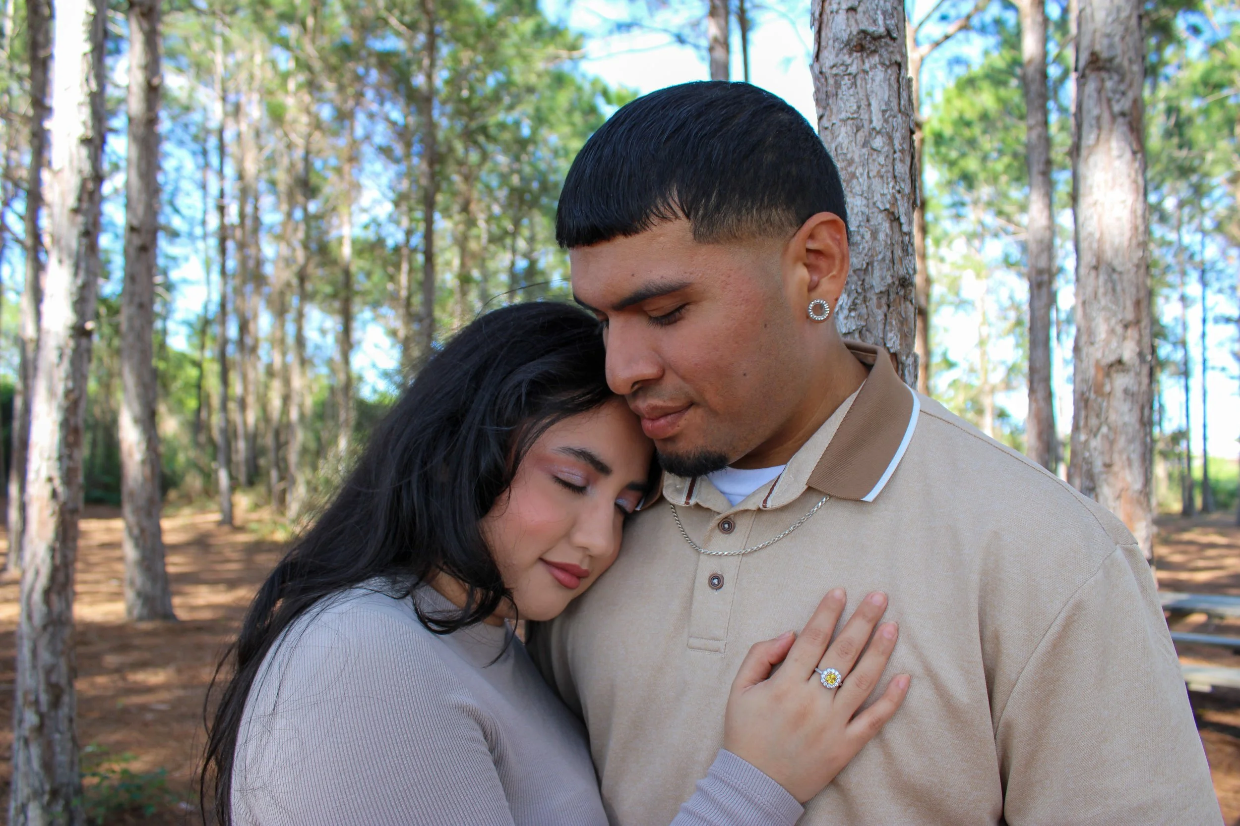 A couple hugging with their eyes closed in a wooded park, the woman resting her head on the man's chest and the man with his eyes closed, wearing earrings and a beige shirt.
