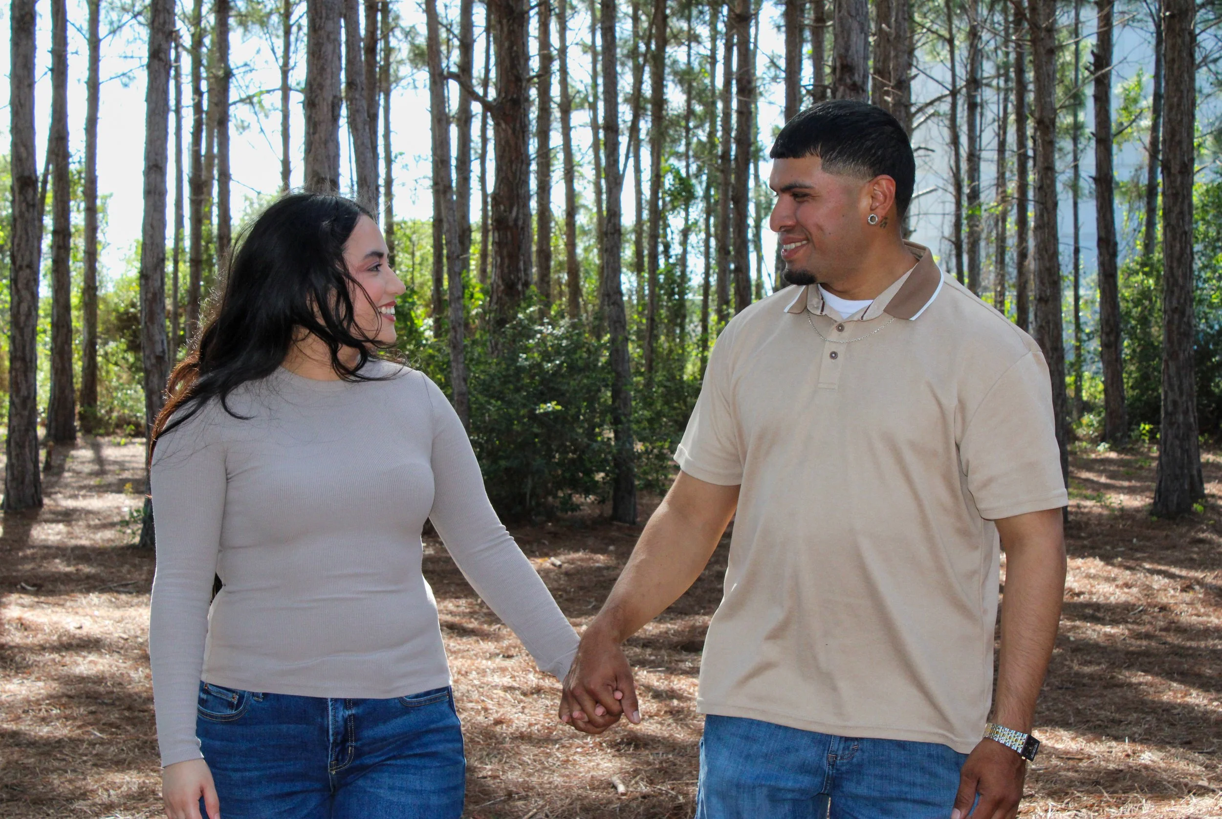 A couple holding hands and smiling at each other while walking in a forest.