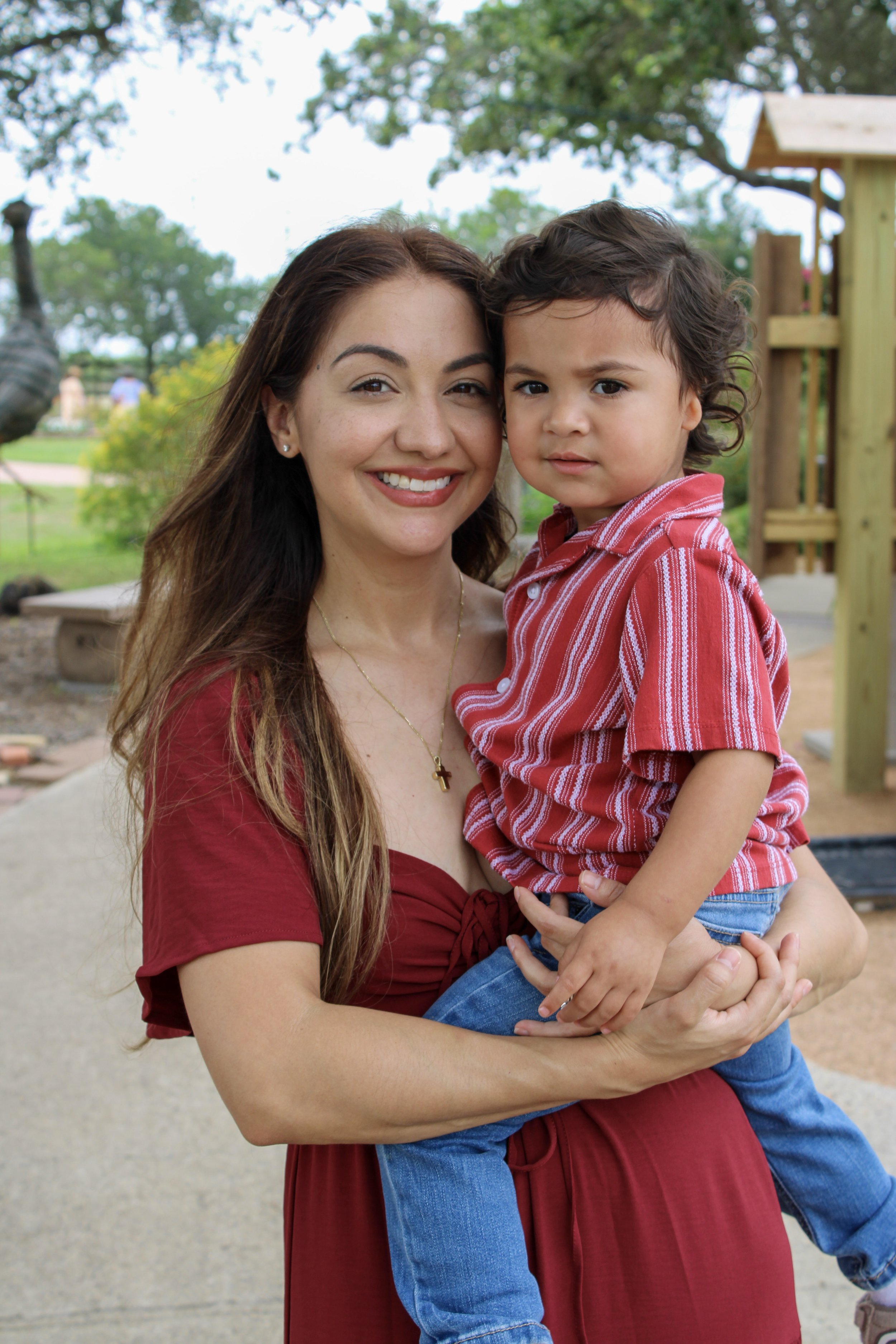 A woman with long brown hair and wearing a red dress is holding a young boy with curly dark hair, dressed in a red and white striped shirt, outdoors in a park with trees and playground equipment in the background.