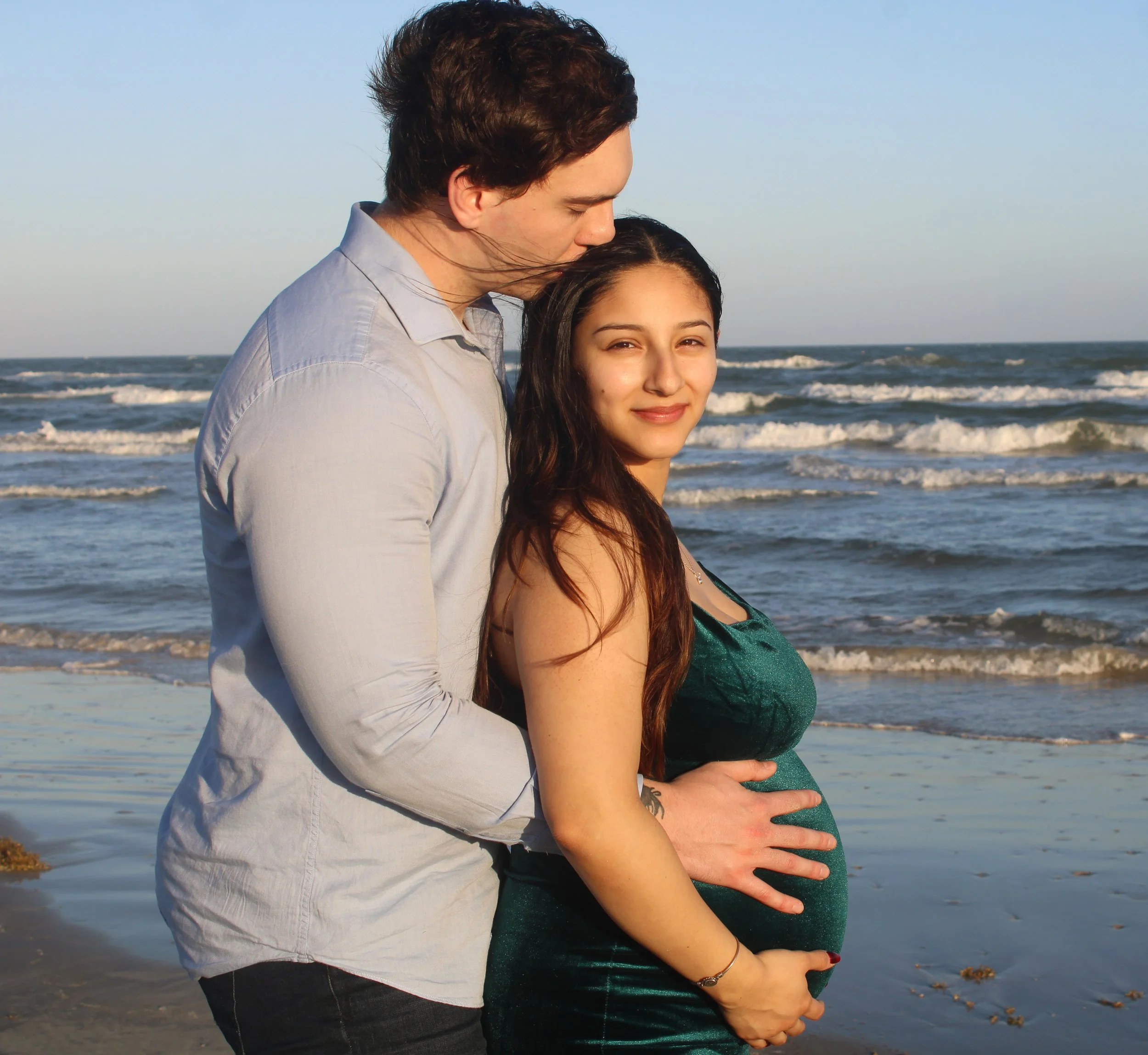 A young couple standing on the beach, with the man gently kissing the woman's forehead. The woman is pregnant, holding her belly, while the ocean waves roll in the background during sunset.