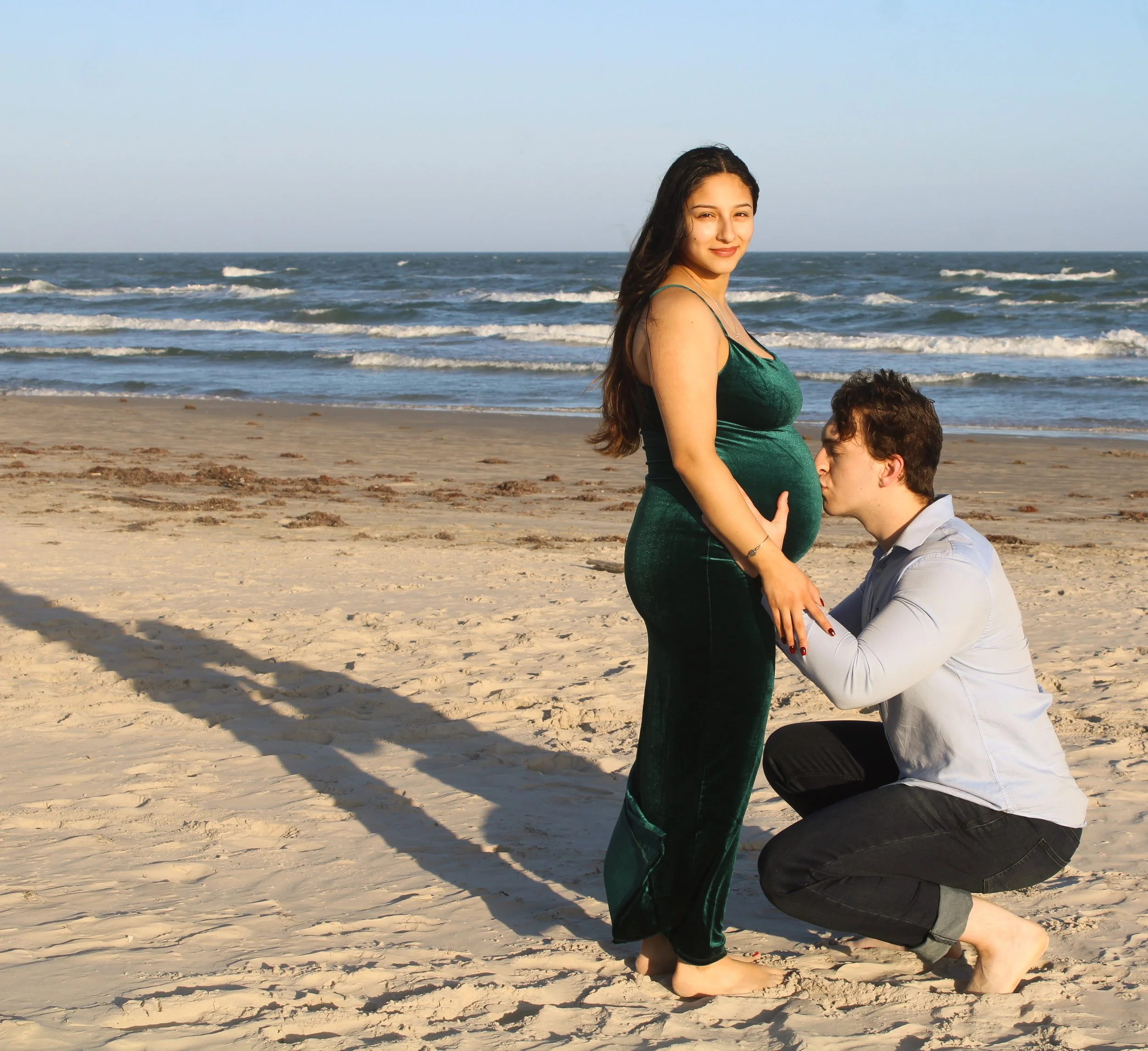 A pregnant woman standing on a sandy beach with ocean waves in the background, smiling at the camera while a man kneels in front of her, kissing her belly.