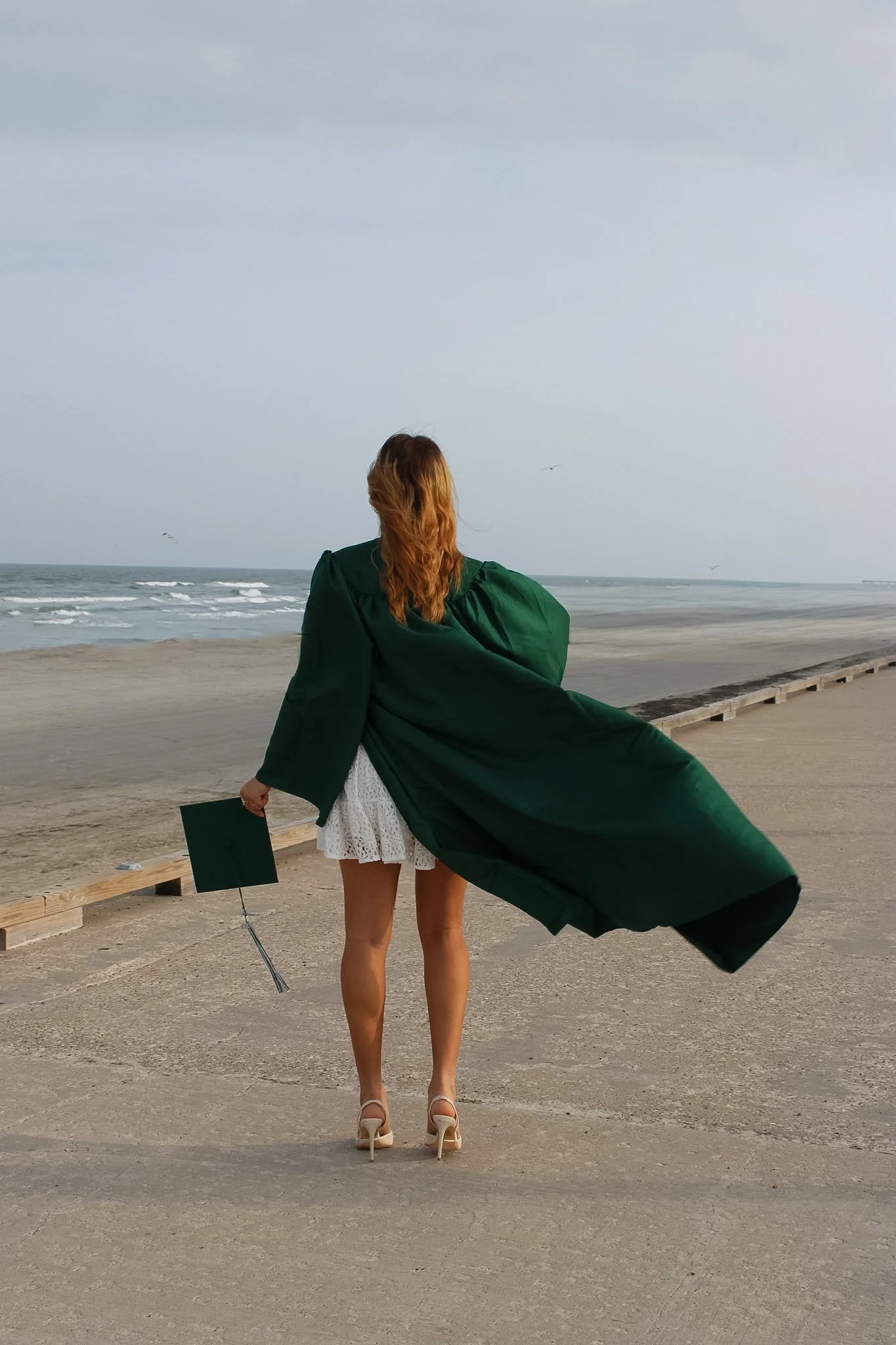 A woman with red hair wearing a dark green coat, white lace dress, and high heels, standing on a beach with the ocean and cloudy sky in the background. She is holding her graduation cap and looking out at the sea.