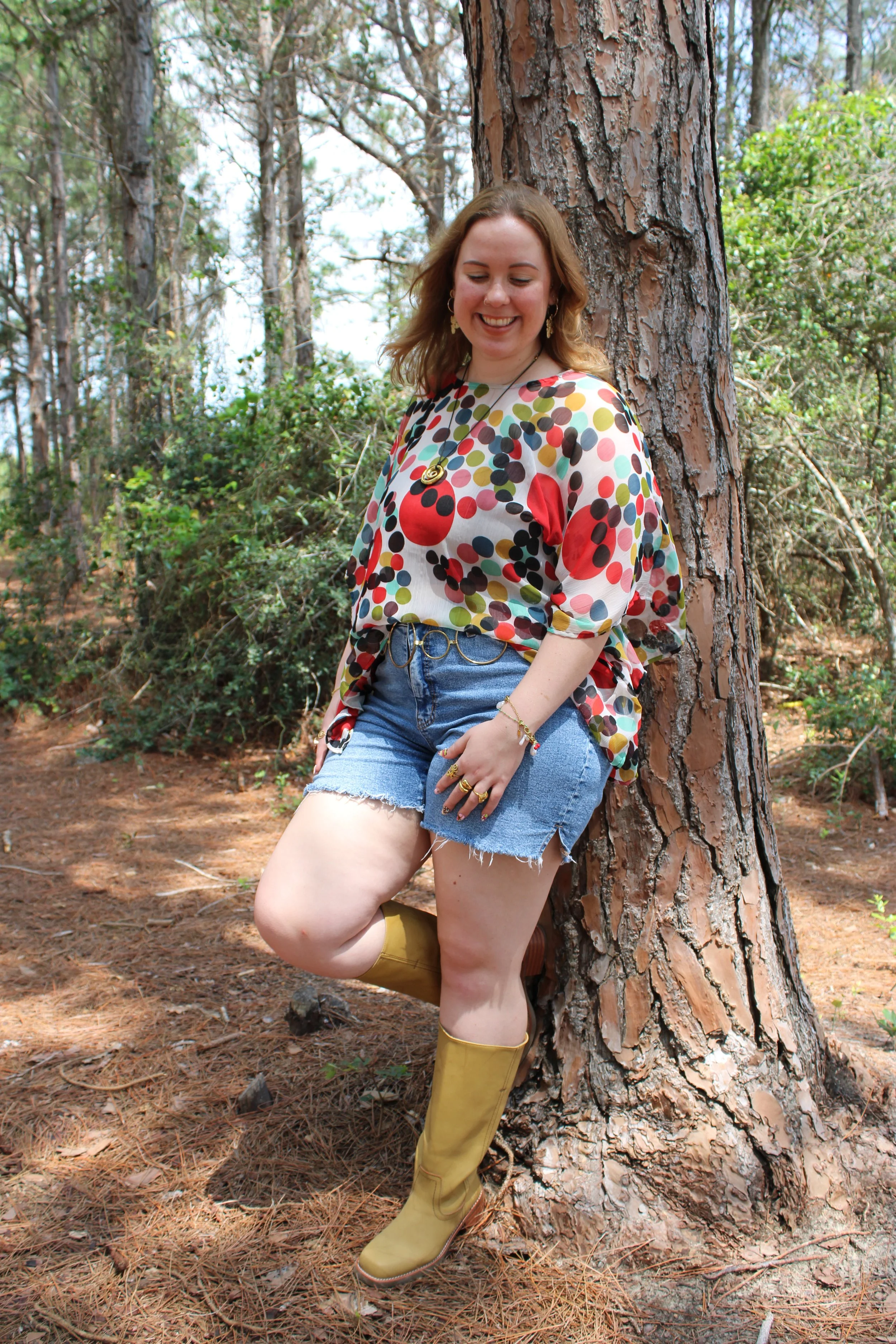 A smiling woman with shoulder-length curly hair wearing a colorful polka dot blouse, denim shorts, and yellow rain boots standing outdoors next to a large tree in a forested area.
