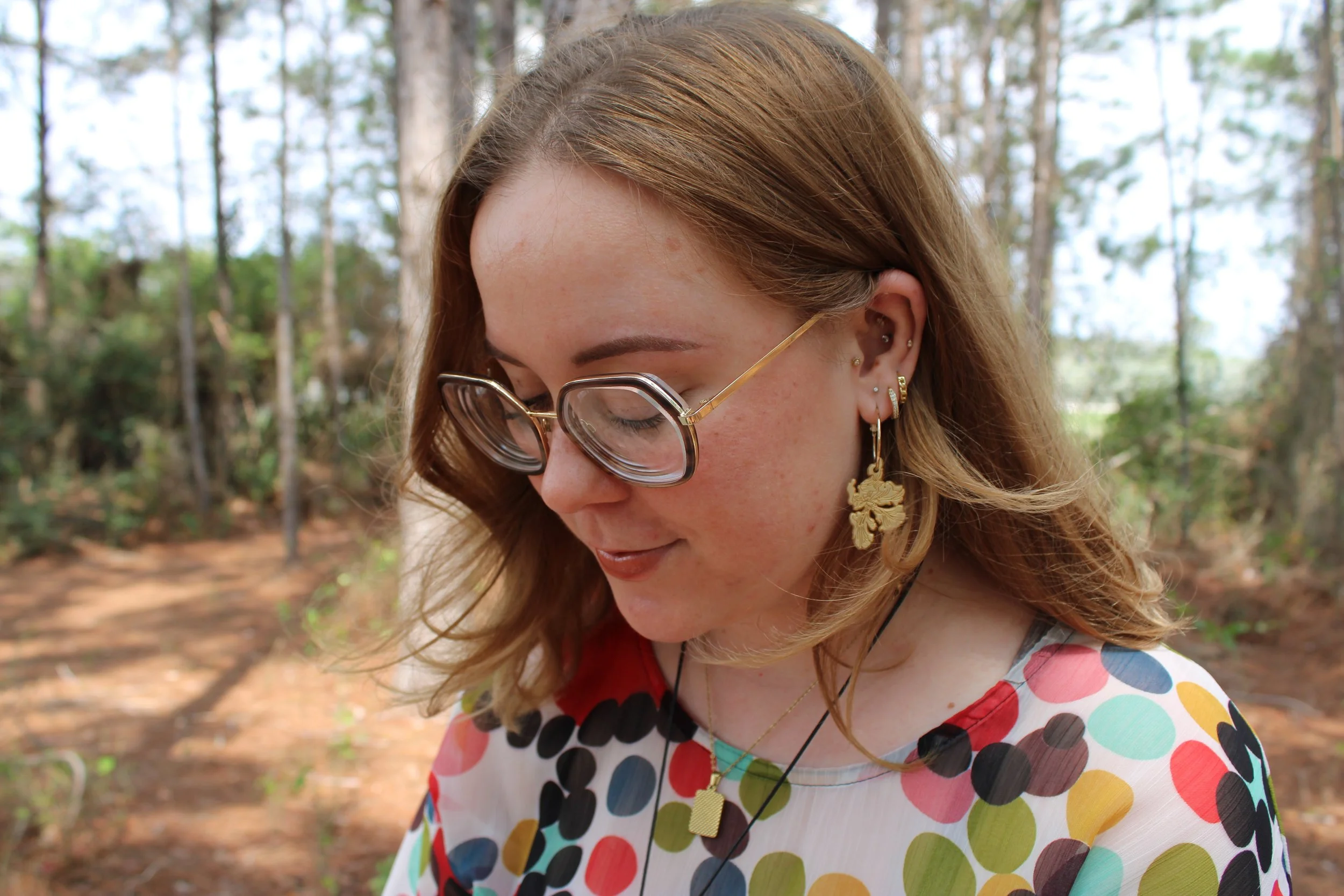 A woman with light brown hair wearing glasses, earrings, and a colorful polka dot top stands outdoors in a wooded area with trees and sunlight filtering through.