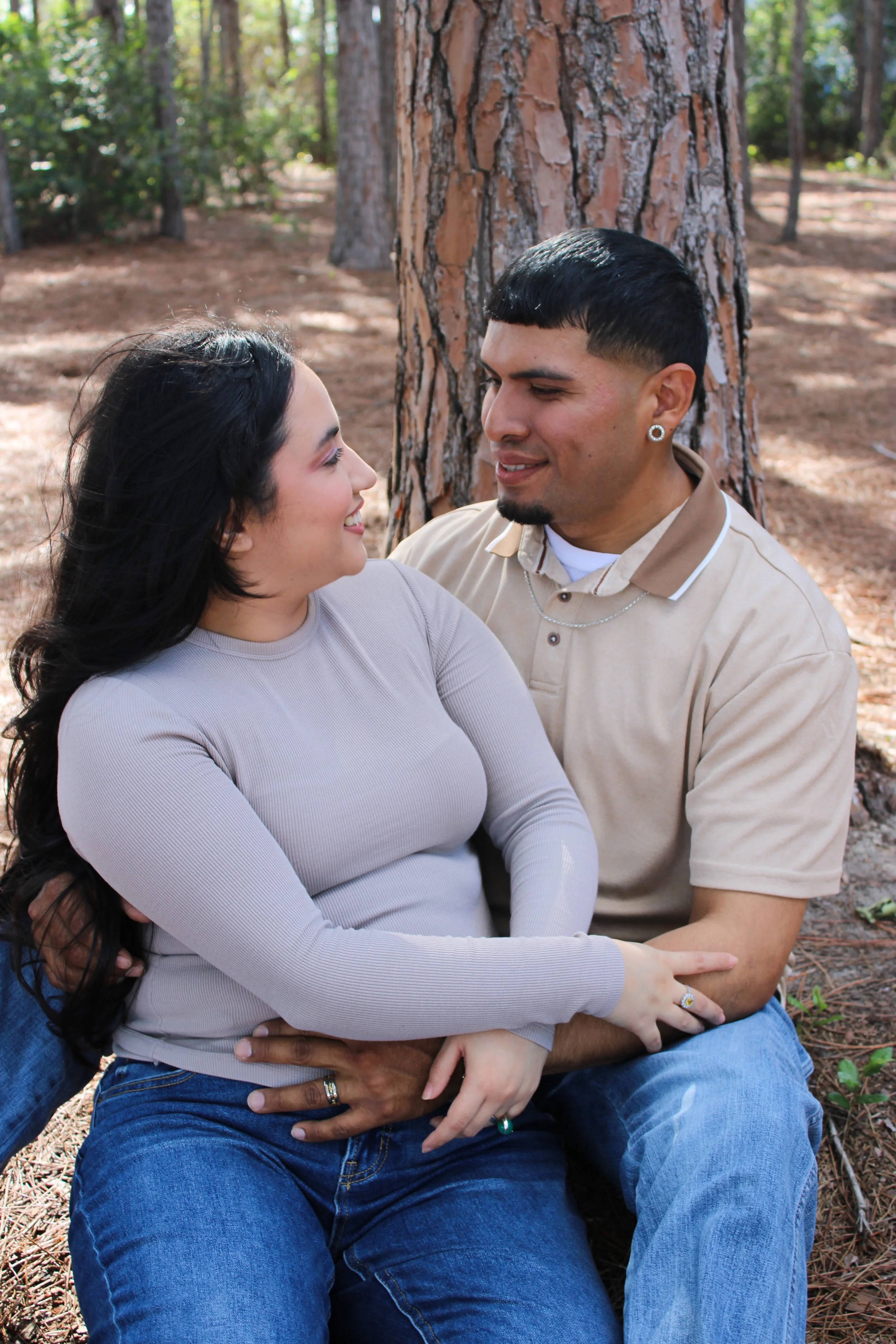 A couple sitting outdoors in a wooded area, embracing and smiling at each other.