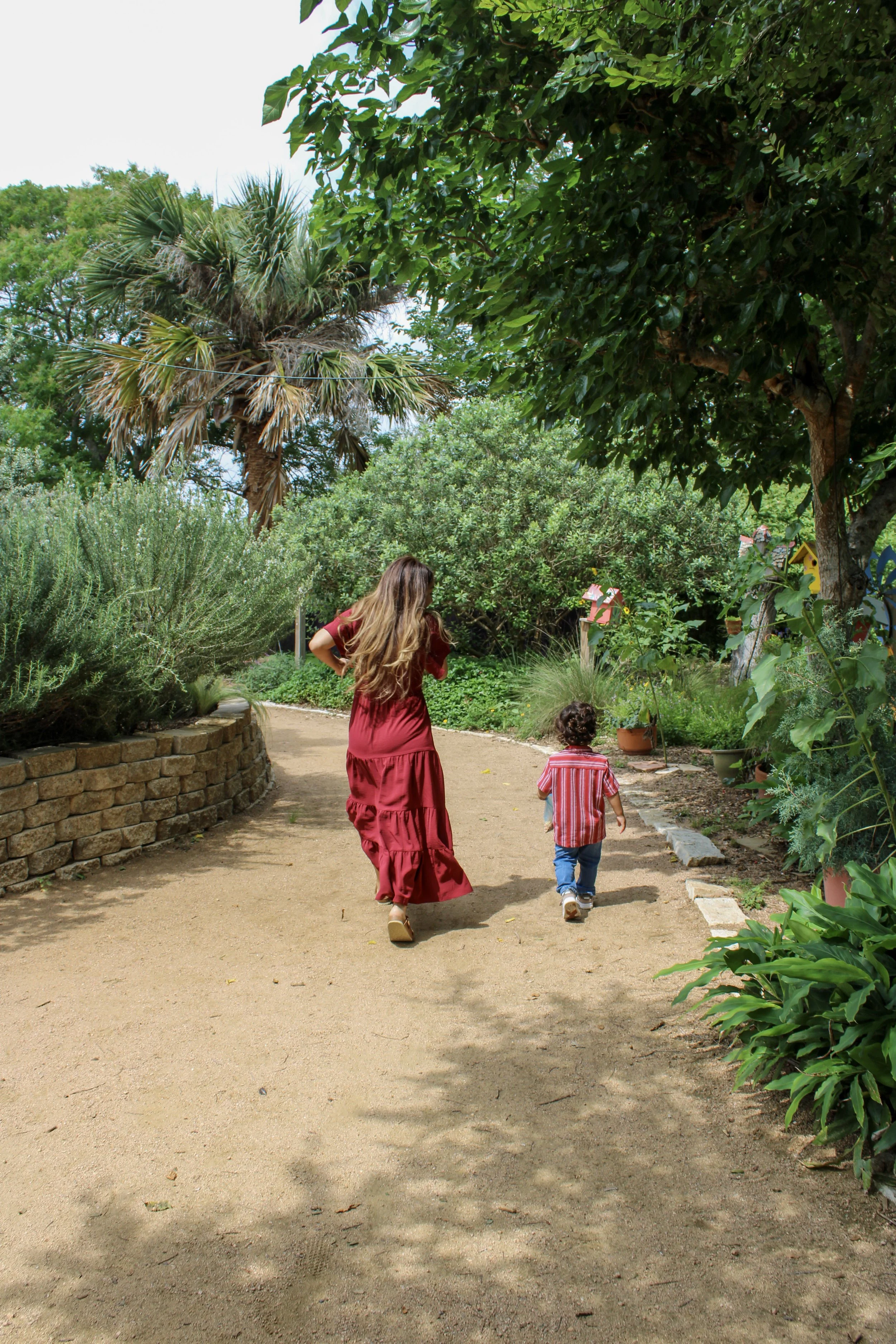 A woman and a young boy walking along a dirt path in a lush garden with various green plants, trees, and birdhouses.
