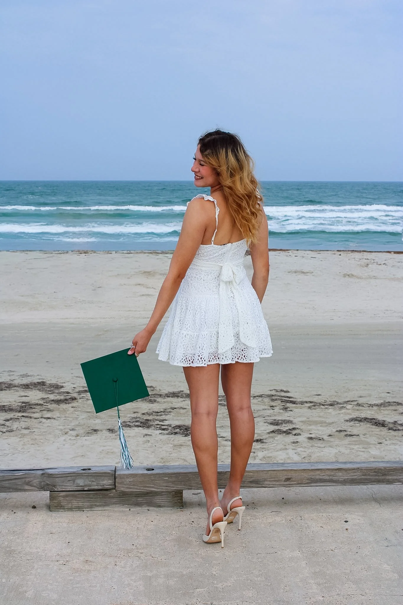 A woman in a white lace dress, smiling with her eyes closed, standing on a beach near the water, holding a green graduation cap in her left hand.
