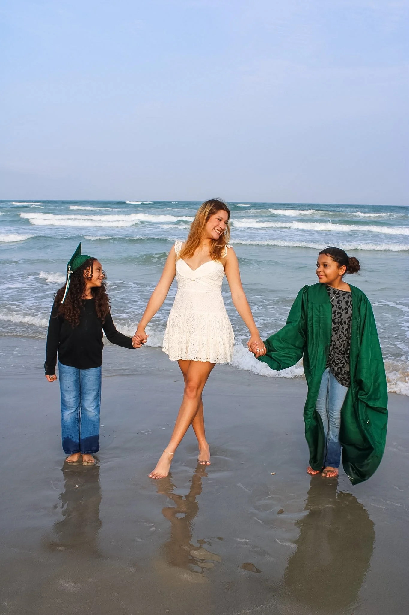 A woman in a white dress holding hands with two young girls, one in a black jacket and blue jeans, the other in a green graduation gown, standing on the beach near the ocean with waves.