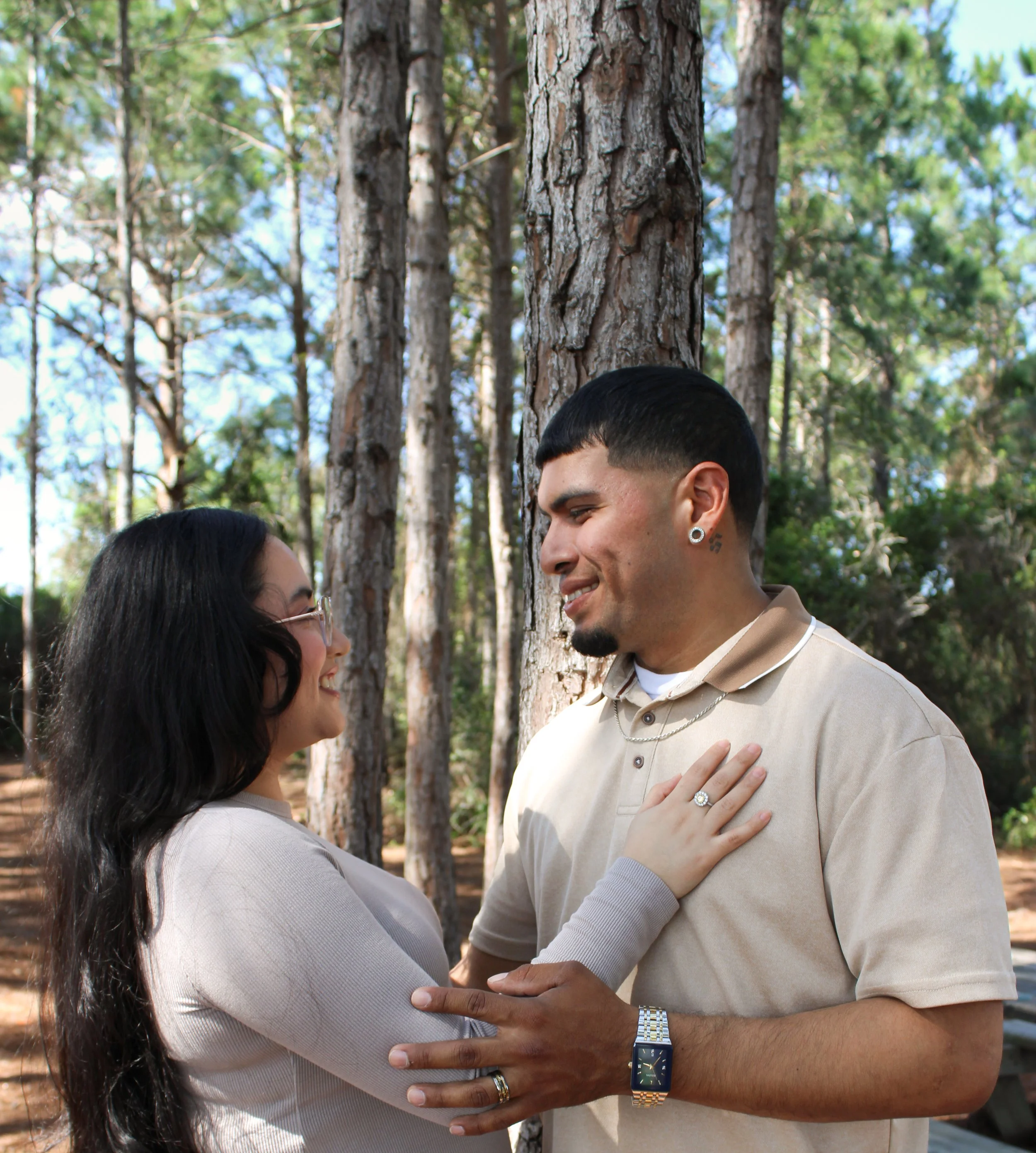 A couple standing close together outdoors in a forest, holding hands and smiling at each other, with trees and sunlight in the background.