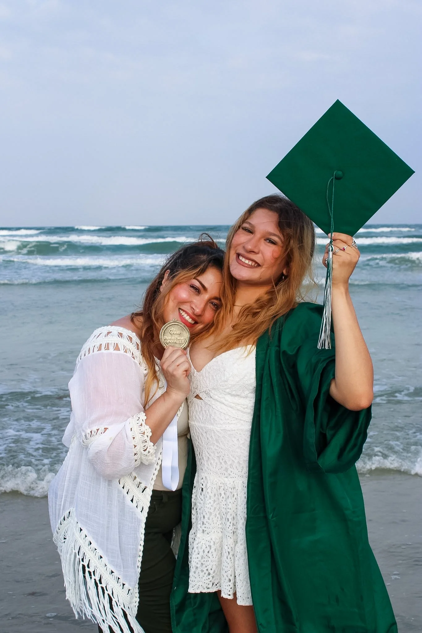 A mother and daughter at the beach, celebrating the daughter's graduation, one in an academic cap and gown holding a diploma, smiling and hugging, with waves in the background.