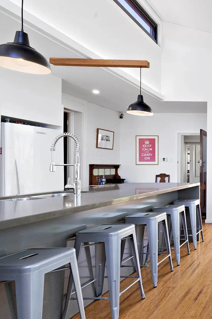 Modern kitchen with a gray island counter, six gray metal stools, black hanging pendant lights, a white refrigerator, a faucet with a flexible sink hose, framed wall art, and wooden flooring.