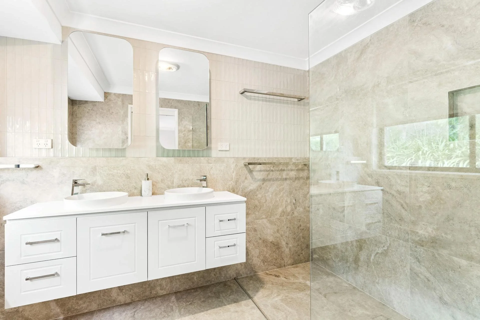 Modern bathroom with white vanity, two sinks, large mirrors, tiled walls and floor, partially visible glass shower with tiled walls and a window.
