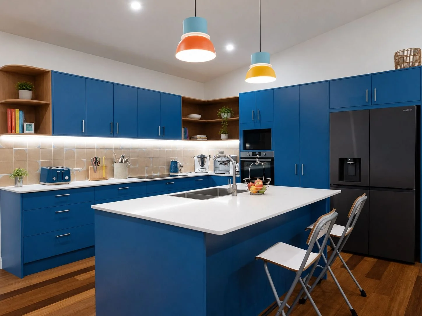 Modern kitchen with blue cabinets, white countertops, and a kitchen island. Equipped with kitchen appliances and utensils. Two chairs are at the island, and colorful pendant lights hang from the ceiling.