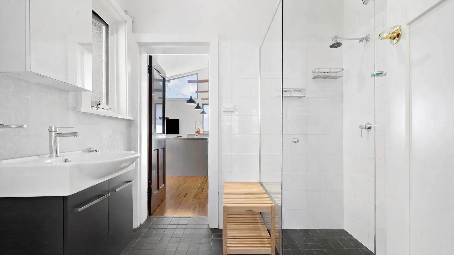 Modern bathroom with white tiled walls, black floor tiles, a white sink with a dark cabinet, a walk-in shower with glass doors, and a wooden bench. An open door leads to a space with pendant lighting and a kitchen area.