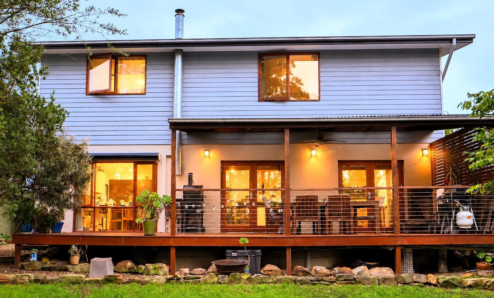 The back view of a two-story house with a large wooden deck. The house has blue siding on the upper level and a beige lower level. The deck features a barbecue grill, dining table, and chairs, lit by outdoor wall lights. The back yard has a grassy la