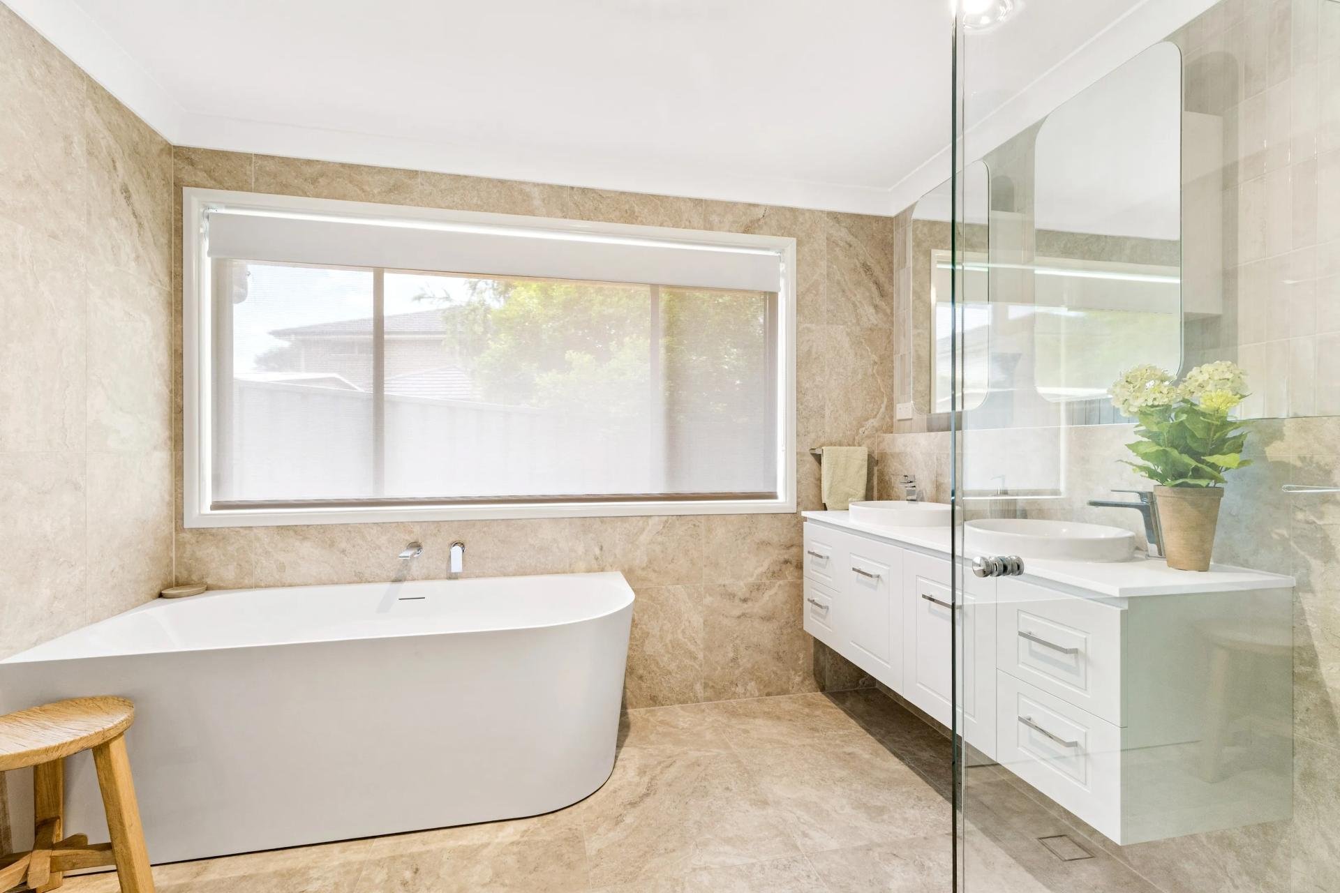Modern bathroom with a large bathtub, white vanity with two sinks, and a glass shower enclosure. There is a potted plant on the vanity and a window with blinds letting in natural light.