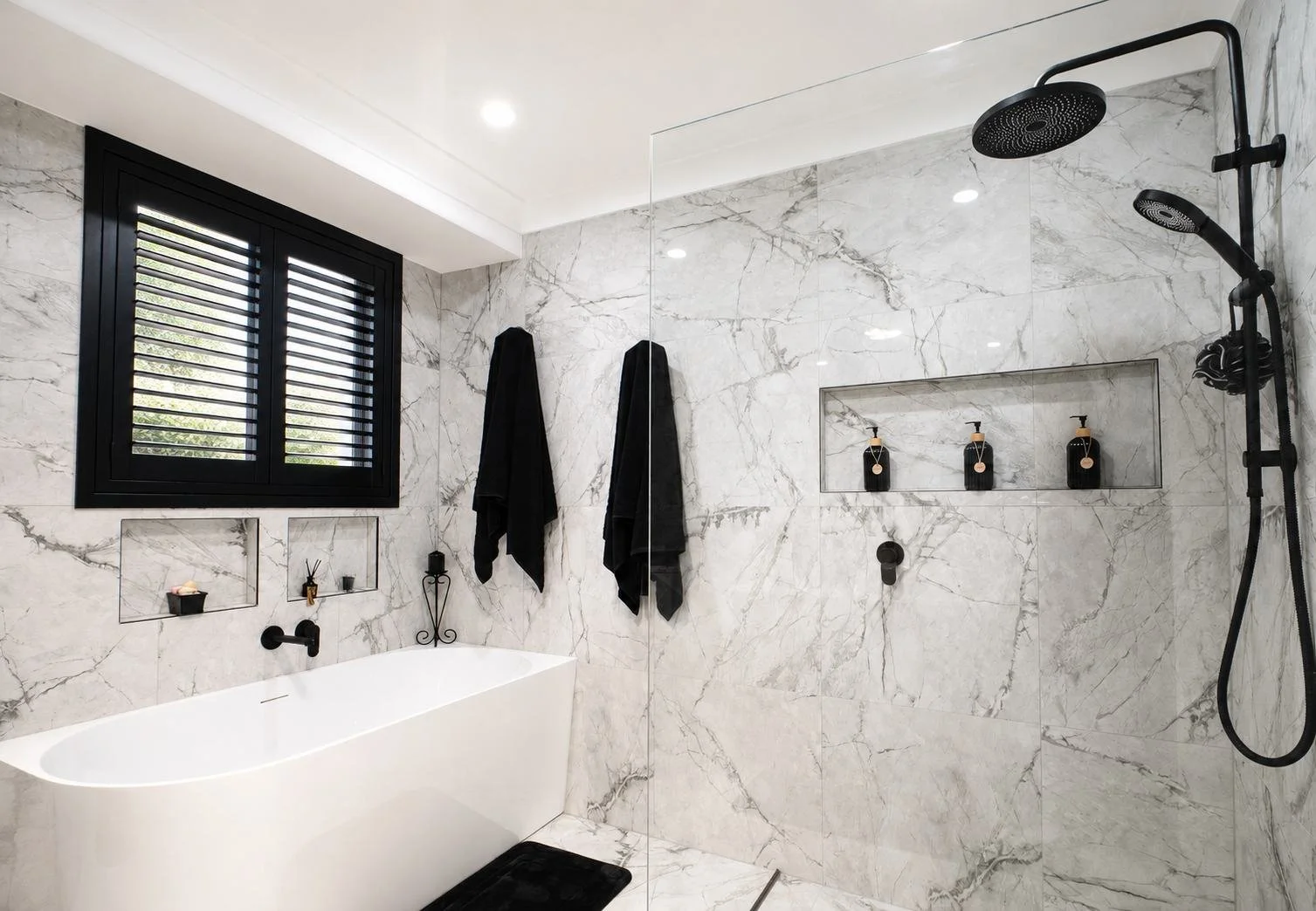 Modern bathroom with marble walls, black window frame with shutters, bathtub, black towels hanging, and a walk-in shower with black shower fixtures and built-in shelves.