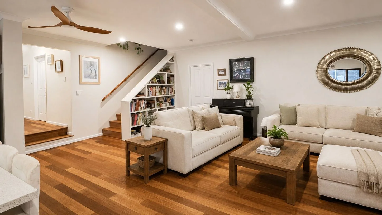 A modern living room with white walls, hardwood floors, and beige sofas. There is a wooden coffee table with a book and plant decor, a side table with a vase, a black piano with plants on top, and a large decorative mirror on the wall. A ceiling fan 