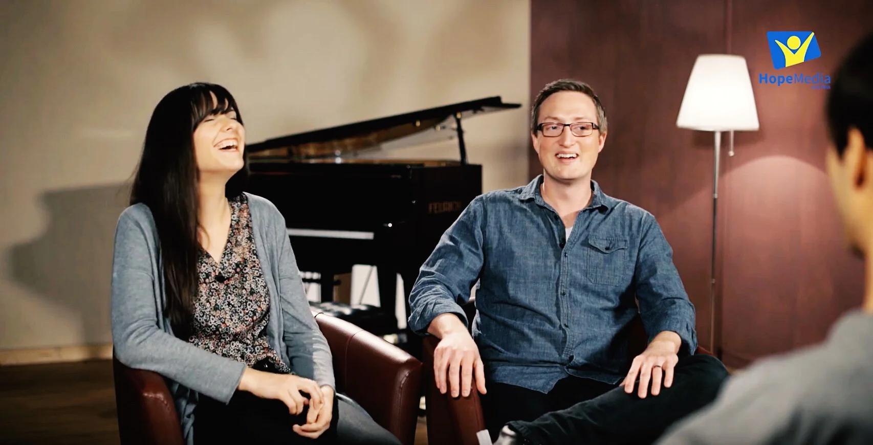 Three people sitting on chairs in a room, laughing and having a conversation, with a grand piano and a floor lamp in the background.