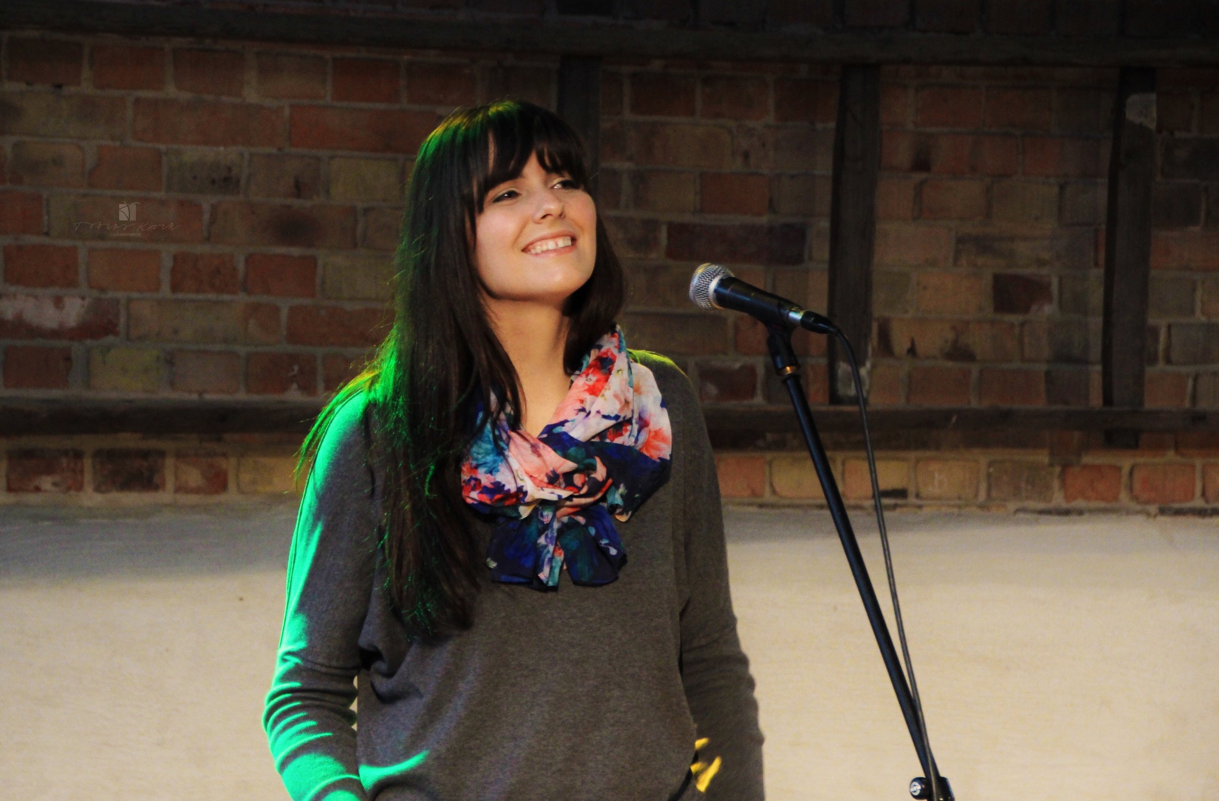 A young woman with long dark hair and bangs, smiling, stands in front of a microphone on stage with a brick wall in the background, wearing a gray sweater and a colorful floral scarf.