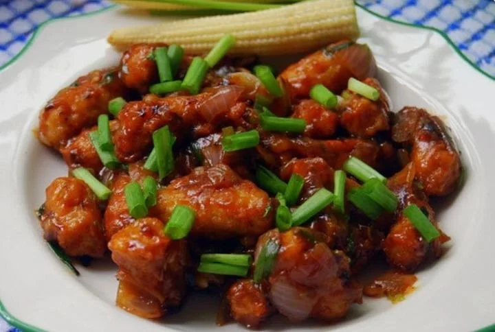 Close-up of a plate with saucy chicken pieces garnished with chopped green onions, accompanied by a slice of cucumber and a piece of baby corn.