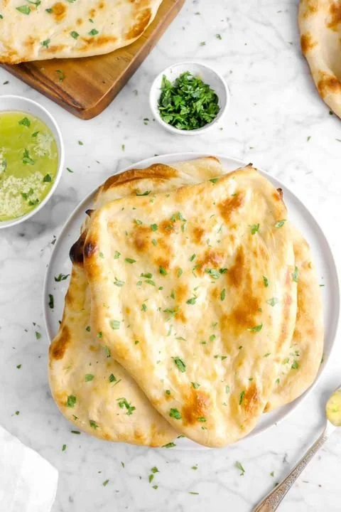Plate of garlic naan bread garnished with chopped cilantro, with small bowls of green chutney and green sauce nearby, on a white marble surface.