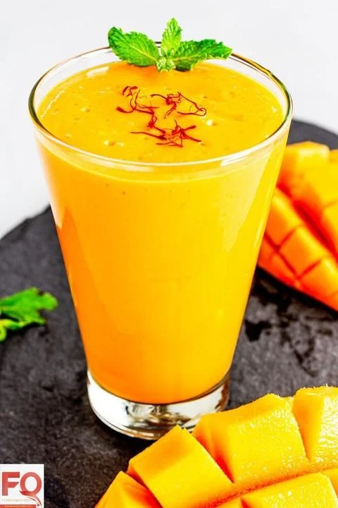 A tall glass of mango smoothie garnished with mint leaves and saffron threads, placed on a black slate serving board with sliced mango pieces nearby.