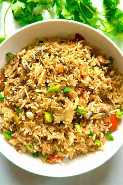 A bowl of cooked rice mixed with vegetables and seasonings, garnished with chopped green onions, and surrounded by fresh cilantro leaves.