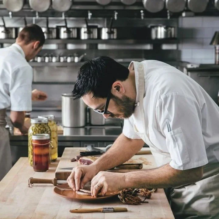 Chef preparing food on a kitchen counter with jars of preserved vegetables in the background.