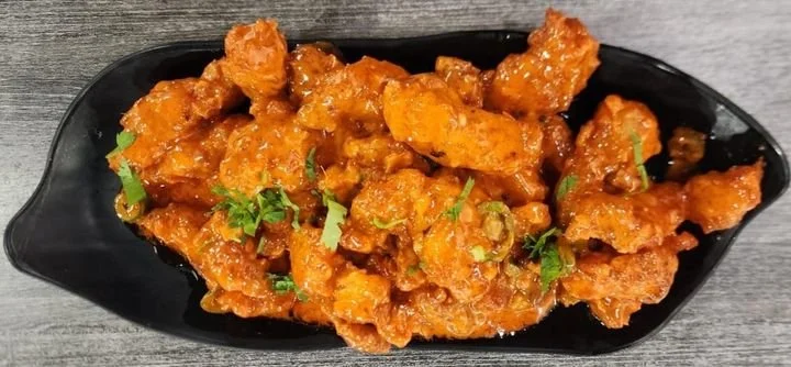 Close-up of crispy orange-colored chicken pieces garnished with chopped green herbs, served on a black plate.