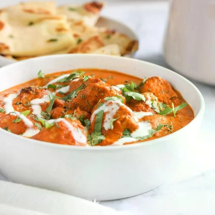 A bowl of Indian butter chicken garnished with chopped cilantro and drizzled with cream, with naan bread in the background.