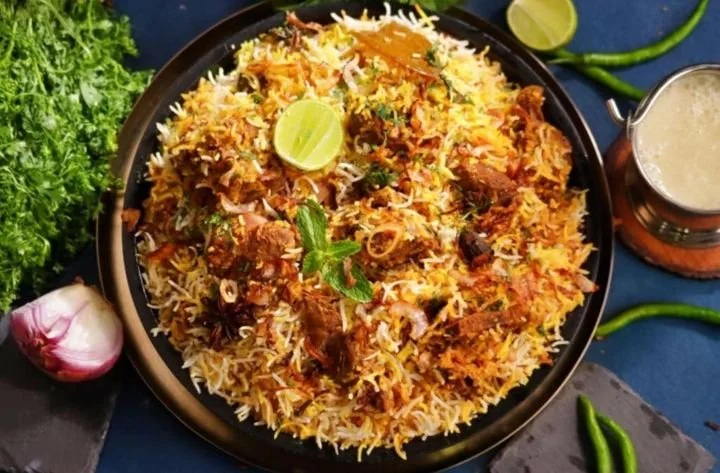A plate of biryani garnished with mint leaves, lemon wedge, and fried onions, accompanied by lime wedges, green chilies, and a small pitcher of raita, with fresh vegetables and garlic nearby.