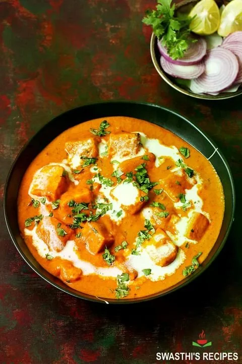 A bowl of Indian butter chicken curry garnished with cilantro, served with sliced onions and lime wedges on a side plate.
