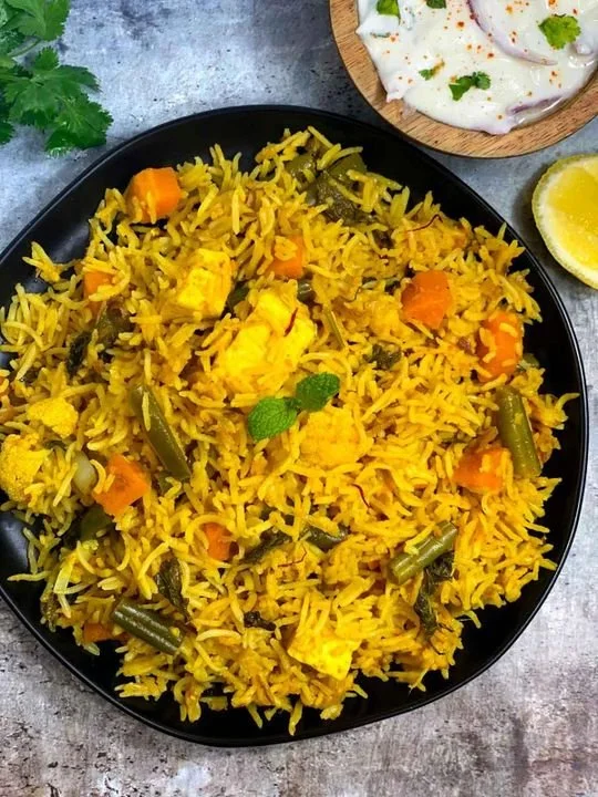 Plate of yellow rice with carrots and green beans, garnished with a sprig of mint, on a gray surface with lemon wedge and side of raita topped with cilantro visible.