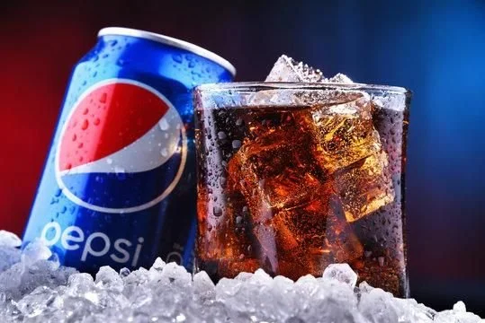 A can of Pepsi next to a glass of cola with ice on ice and a dark background.