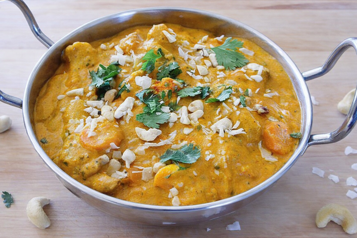 A metal bowl filled with curry topped with cashews and cilantro, placed on a wooden surface.