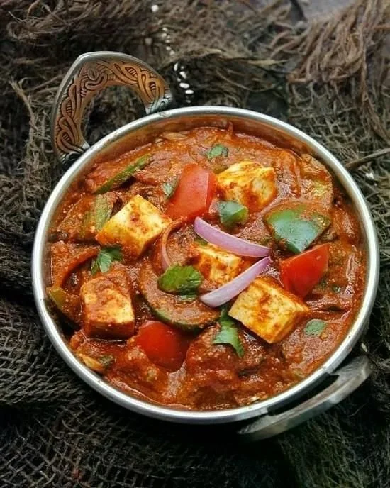 A bowl of Indian vegetable curry with tofu, tomatoes, zucchini, and onions, garnished with cilantro, in a silver bowl with a decorative handle on a rustic dark surface.