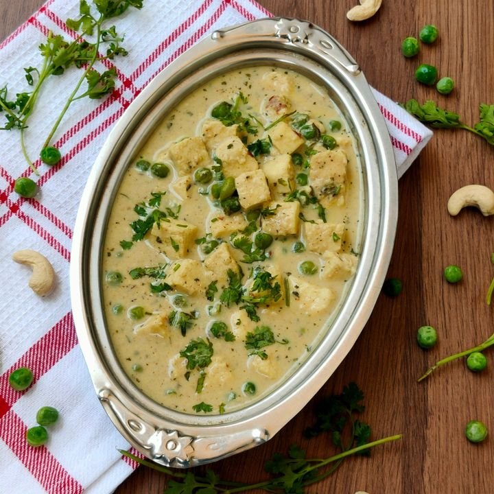 A dish of vegetable curry with green peas, tofu, and cilantro in a creamy sauce served in a silver bowl on a wooden table with scattered green peas, cashews, and cilantro leaves.