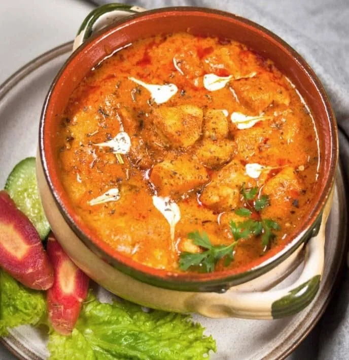 A bowl of Indian chicken curry garnished with fresh herbs, served with sliced cucumbers and red radishes on a side plate.