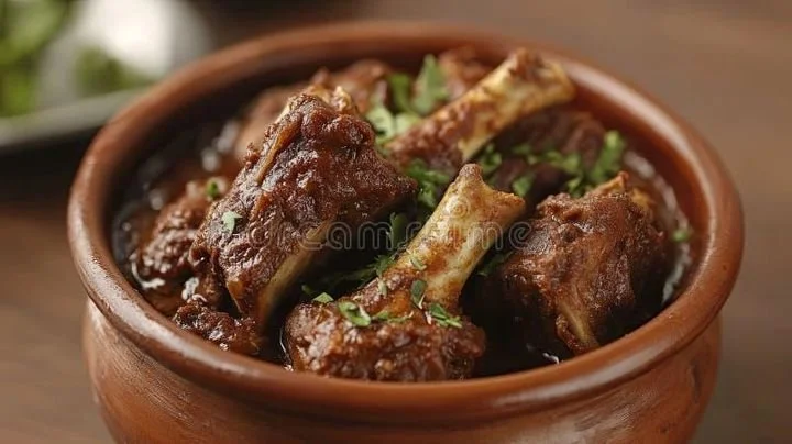 A small clay bowl of spicy beef stew with chunks of meat and bones garnished with chopped green herbs.