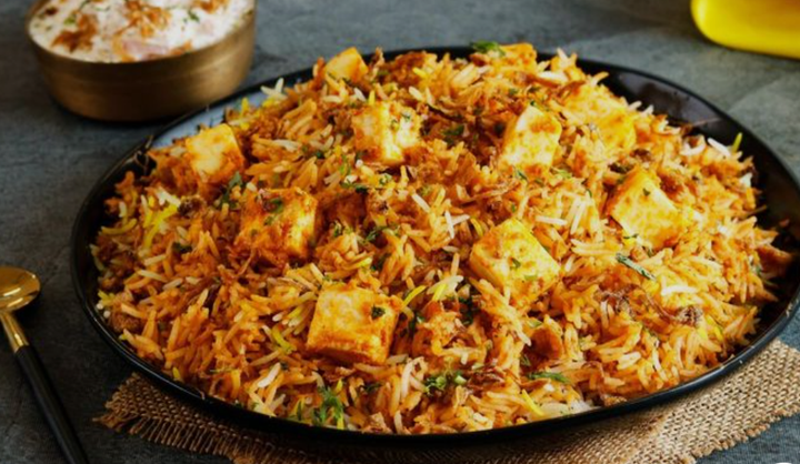 A black oval dish filled with chicken biryani rice topped with herbs, accompanied by a small bowl of raita and a spoon on the side.
