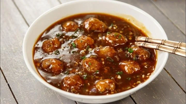 White bowl filled with sweet and sour meatballs in a dark sauce on a gray wooden surface with a silver utensil resting on the bowl's edge.