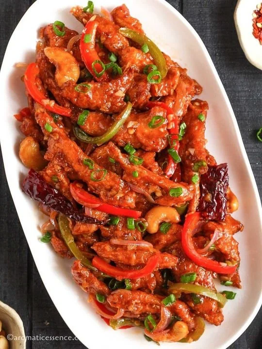 Stir-fried dish with chunks of meat, red and green bell peppers, cashews, and garnished with chopped green onions on a white oval plate