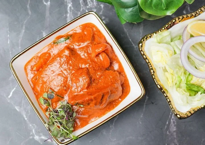 A dish of chicken in a red sauce served in a square white bowl with gold trim. Next to it is a bowl of chopped lettuce and sliced onions. The bowls are on a dark marble surface and there are green leaves in the background.