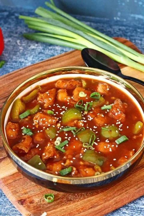 A bowl of spicy Asian-style chicken stew with green onions and sesame seeds, on a wooden cutting board, with fresh green onions in the background.