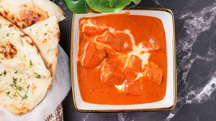 A bowl of Indian butter chicken curry with chunks of chicken in a creamy tomato sauce, garnished with a small green leafy herb, next to naan bread.