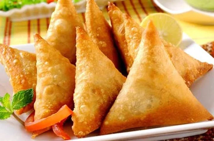 Golden-fried samosas on a white platter with lime wedges and green garnishes.